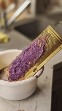 A kitchen grater with which purple potatoes are grated into a white bowl to make dumpling dough,