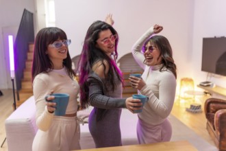 Three happy young women friends wearing sunglasses and casual clothes dancing, holding mugs, and