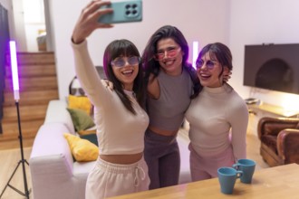 Three cheerful young women wearing colorful sunglasses gathering for a selfie in a modern living