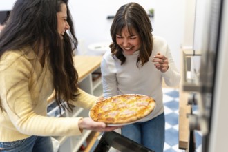 Two cheerful friends laugh in a cozy kitchen while presenting a freshly baked homemade pizza,