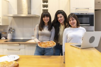 Three young women smiling and looking at camera while one is holding a freshly baked pizza in a