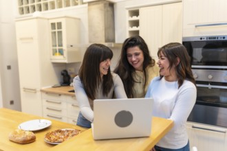 Three young women laughing together in a bright modern kitchen, watching a laptop while sharing