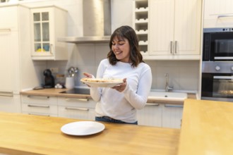 Young woman smiling in a modern kitchen while holding a freshly baked pizza on a plate, enjoying