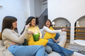 Three women on a sofa sharing popcorn, laughing and watching tv together in a cozy living room,