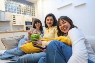 Three young women laughing and relaxed on a couch at home, enjoying a cozy movie night together