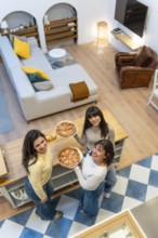 Three cheerful young women standing in an open plan apartment, holding pizzas and looking up while