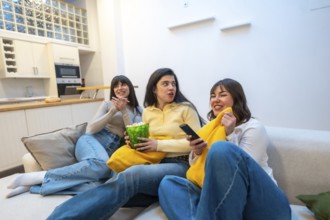 Three casual women friends relaxing on a comfortable sofa, watching a movie, eating popcorn,