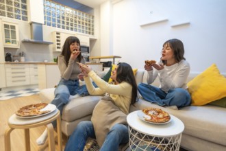 Three smiling young women relax on a modern sofa in an apartment, sharing pizza and laughter during