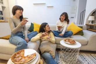 Three young women laughing and talking while eating pizza slices on a sofa in a modern living room,