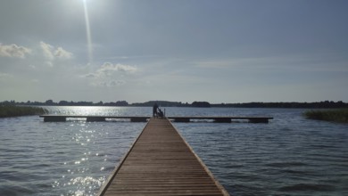 A long wooden walkway leads across a calm lake under bright sunlight, Masuria, Poland