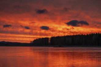 A dramatic sunset over a still, frozen lake and forest with orange-red hues in a cloudy sky,