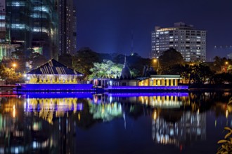 At night, a brightly lit temple is reflected in the calm waters of an urban lake, The Seema Malaka