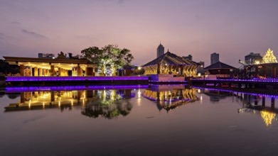 An illuminated temple and city building are reflected in the tranquil lake at dusk, The Seema