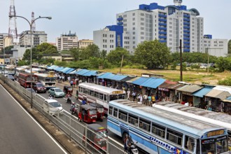 City street with several buses and market stalls, surrounded by large buildings and trees, buses in