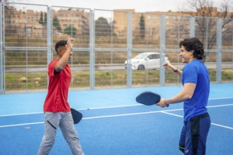 Two men in red and blue shirts smile and high five on a vibrant blue outdoor pickleball court,
