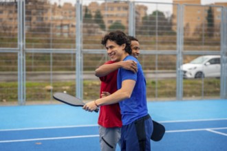 Two diverse male friends smiling and hugging on a blue pickleball court after a game, demonstrating