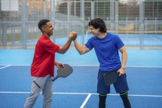Two diverse men playing pickleball are celebrating with a high five on a blue outdoor court,