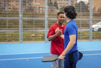 Two young multi ethnic men shaking hands and smiling on a blue pickleball court after a match,