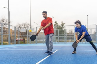 Two young men play pickleball on a bright blue outdoor court, one about to hit a yellow ball with