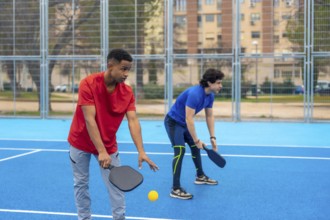 Young men poised on a bright blue court with paddles and ball, ready for an outdoor pickleball