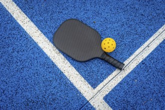 Pickleball paddle and yellow wiffle ball resting on the vibrant blue court surface with distinct