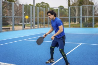 Young man playing pickleball on a blue outdoor court, striking a yellow ball with a black paddle,