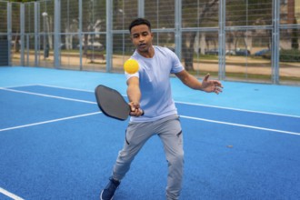 Young man strikes a yellow pickleball with a paddle on a vibrant blue outdoor court, showcasing
