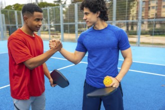 Two young diverse male athletes shaking hands on a blue pickleball court, holding paddles and a