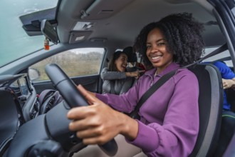 Smiling young black woman driving a car with friends in the back, laughing and pointing, capturing