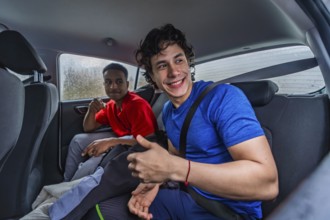 Two young diverse men sitting in the back seat of a car, one smiling and giving a thumbs up,