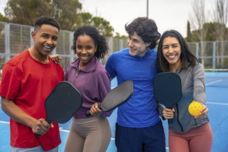 Group of diverse young friends smiling and embracing, holding pickleball paddles and a ball on a