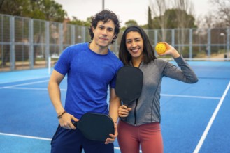 Enthusiastic young couple standing on a blue pickleball court, smiling and holding paddles and a