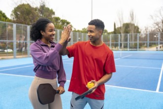 Young diverse couple playing pickleball on a blue court, celebrating with a high five, showing
