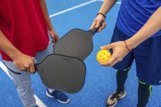 Two people are ready to play pickleball, holding paddles and a bright yellow wiffle ball on a
