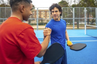 Two diverse male pickleball players shaking hands on a blue court, proudly displaying their paddles