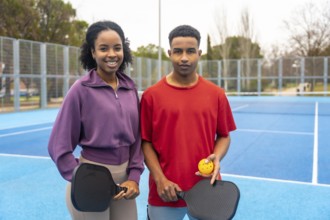Young couple standing on a blue pickleball court, holding paddles and a ball, smiling at the