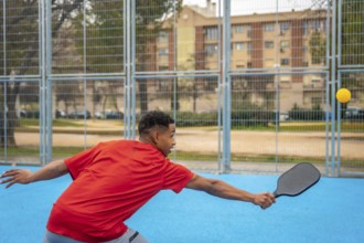 Young man in a red top lunging with paddle to hit a yellow pickleball on a bright blue outdoor