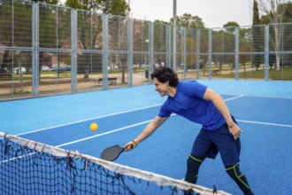 Young man in sports clothing enthusiastically playing pickleball on a bright blue court, hitting a