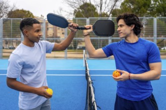 Two men stand on an outdoor pickleball court holding paddles and balls, sharing a friendly moment