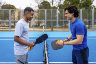 Two diverse young men standing on a vibrant blue pickleball court, holding paddles and balls,
