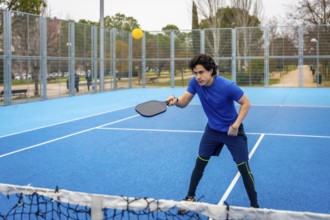 Young man in blue activewear playing pickleball on a blue court, intently hitting a yellow ball