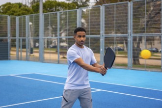 Young man poised to strike a yellow ball with a paddle on an outdoor blue pickleball court, focused
