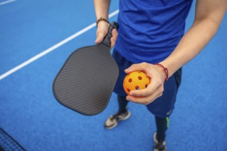 Player standing on a vibrant blue pickleball court, holding a textured paddle and an orange wiffle