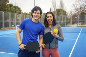 Two young athletes smiling on a bright blue outdoor pickleball court, holding paddles and a ball,