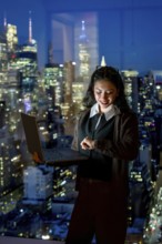 Young businesswoman standing by a large window, working on a laptop and checking her smartwatch