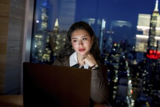 Young businesswoman working late on a laptop in a modern office, gazing toward the illuminated