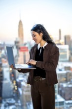 Young businesswoman standing by a window, focused on her laptop while working in an urban setting