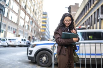 Young executive woman is holding a modern tablet and smiling, standing outdoors in a bustling city