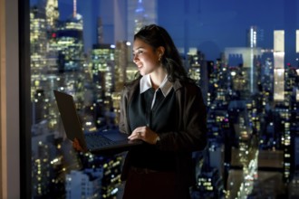 Young businesswoman stands by a floor to ceiling window at night, holding a laptop and smiling at