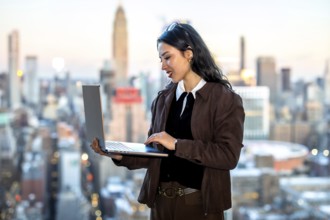 Young businesswoman working on a laptop, standing with a new york city skyline as a blurred
